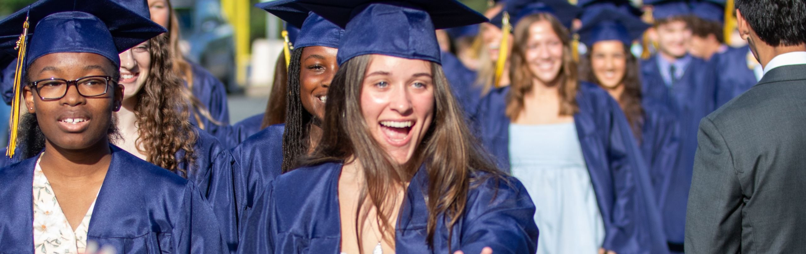 A female graduate in a cap and gown reaches out toward someone on the other side of the camera
